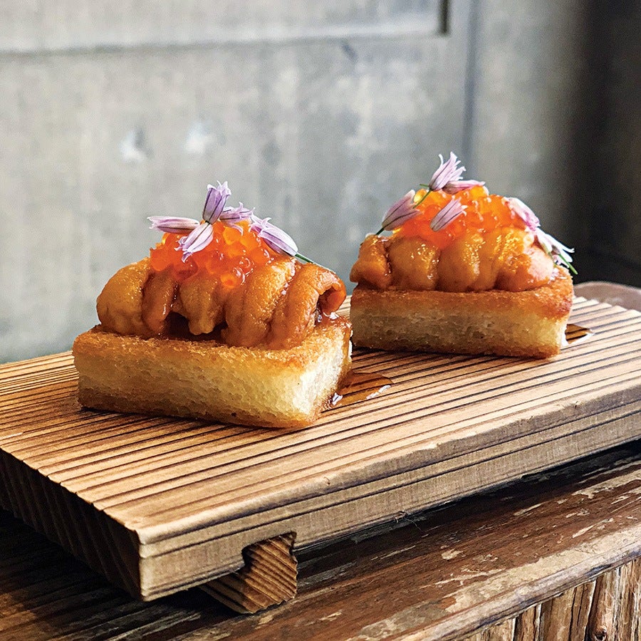 Two pieces of sushi featuring sea urchin (uni) topped with orange fish roe and small purple edible flowers, served on rectangular toasted rice blocks on a wooden board.
