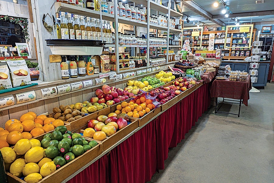 A market display featuring a variety of fresh fruits arranged in wooden crates, including lemons, limes, avocados, oranges, apples, and pears. Above the fruit, shelves hold bottles of juices, sauces, and various packaged goods. The display is covered with a red skirt, and additional tables with baked goods and other products are visible in the background. The setting appears to be a rustic indoor market or store with wooden shelves and a concrete floor.