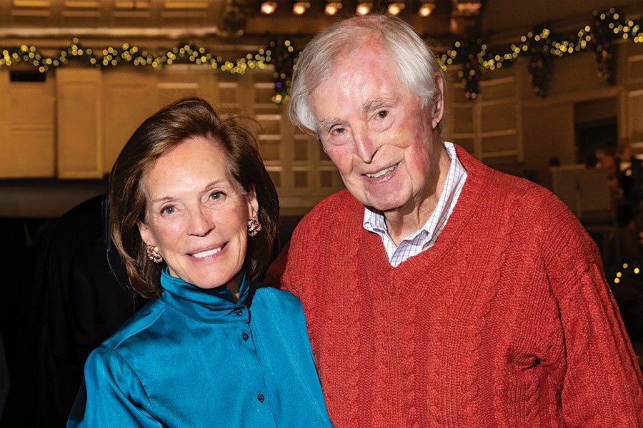 A smiling elderly man wearing a red cable-knit sweater over a checkered shirt stands next to a smiling woman in a shiny teal blouse with button details and decorative earrings. The background features warm lighting and festive garlands with small yellow lights.