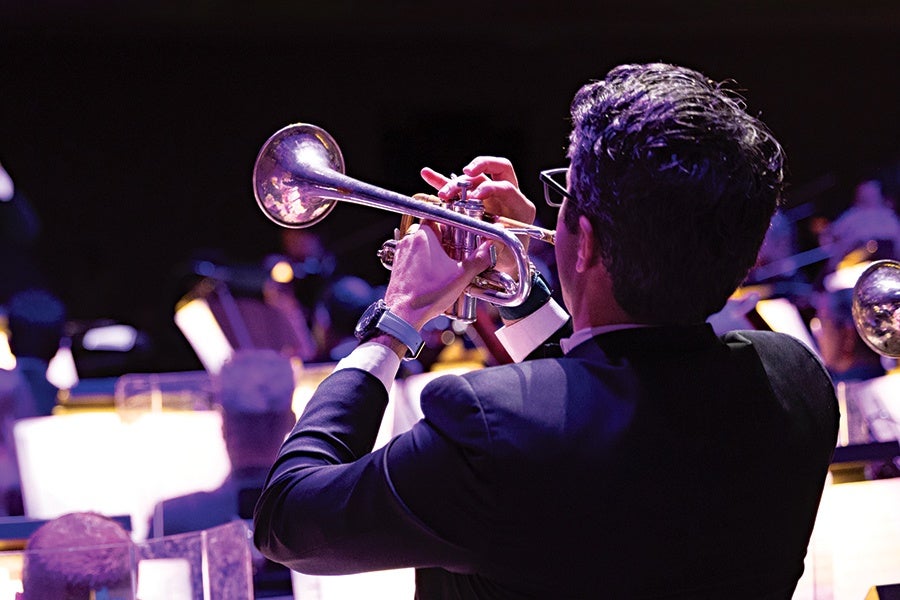 A musician in a dark suit playing a trumpet, viewed from behind, with a blurred orchestra and music stands in the background.