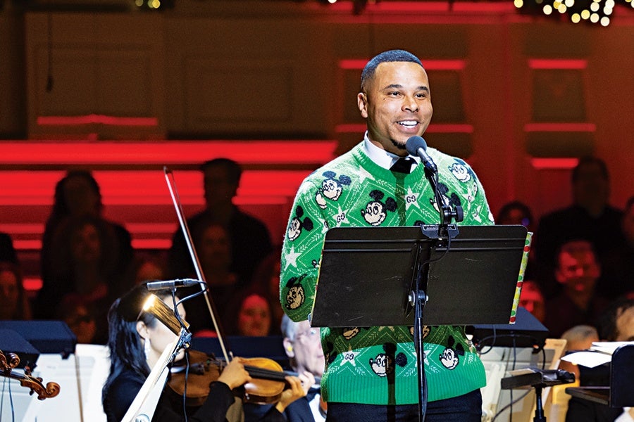 A man wearing a green sweater with Mickey Mouse faces is standing and speaking into a microphone on stage, with musicians playing string instruments in the background. The setting appears to be a concert or performance with red lighting.