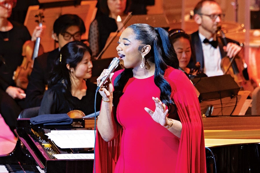 A woman in a bright red dress with long sleeves is singing into a microphone. She has long black hair styled in a high ponytail and is wearing dangling earrings. Behind her, several musicians are playing string instruments, including violins and a cello, and are dressed in formal black attire. A grand piano with sheet music is visible in the foreground.