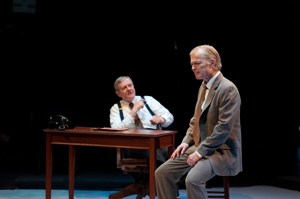 Two men in suits are engaged in a serious conversation on a stage set. One man is sitting on a stool in the foreground, looking contemplative, while the other man is seated behind a wooden desk with a black rotary phone, appearing to speak or gesture. The background is dark, emphasizing the focused lighting on the men and the desk.