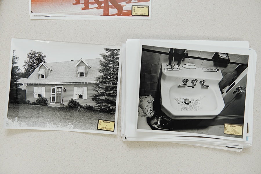 Black and white photographs laid out on a light surface, one showing a house with a lawn and trees, and the other showing a bathroom sink with a faucet, soap dish, and a towel nearby. Both photos have yellow evidence tags labeled "EXHIBIT" with numbers.