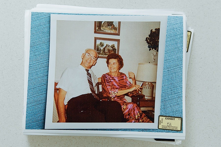 A vintage photograph of an elderly man and woman sitting together indoors. The man is wearing glasses, a white short-sleeve shirt, and a striped tie, while the woman is dressed in a colorful patterned dress. Behind them are two framed paintings on the wall and a table with a lamp and a decorative plant. The photo is placed on top of a stack of papers with a blue textured border and a small label marked "EXHIBIT 51" in the bottom right corner.