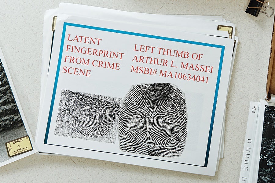 A document labeled "LATENT FINGERPRINT FROM CRIME SCENE" and "LEFT THUMB OF ARTHUR L. MASSEI MSBI# MA10634041" featuring two fingerprint images, one smaller and one larger, both showing detailed ridge patterns. The document is placed on a light-colored surface with other papers and a binder clip nearby.
