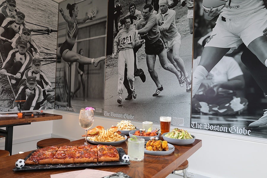 A wooden table displays a variety of food and drinks, including a rectangular pizza, a plate of French fries with a sandwich, a bowl of salad, a plate of roasted corn, a bowl of chicken wings, and a bowl of popcorn. There are also three drinks: a glass of beer, a cocktail with a lime garnish, and a large wine glass with a pink flower decoration. The background features large black-and-white sports photographs from The Boston Globe, depicting rowing, gymnastics, running, and basketball.