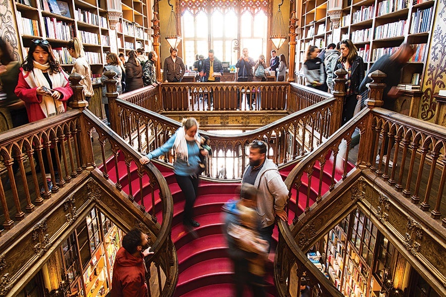 A grand, ornate wooden staircase with red carpet in a library filled with bookshelves. People are walking up and down the stairs and browsing the books. The space is well-lit by a large window at the back, and the walls are decorated with intricate woodwork and patterns.
