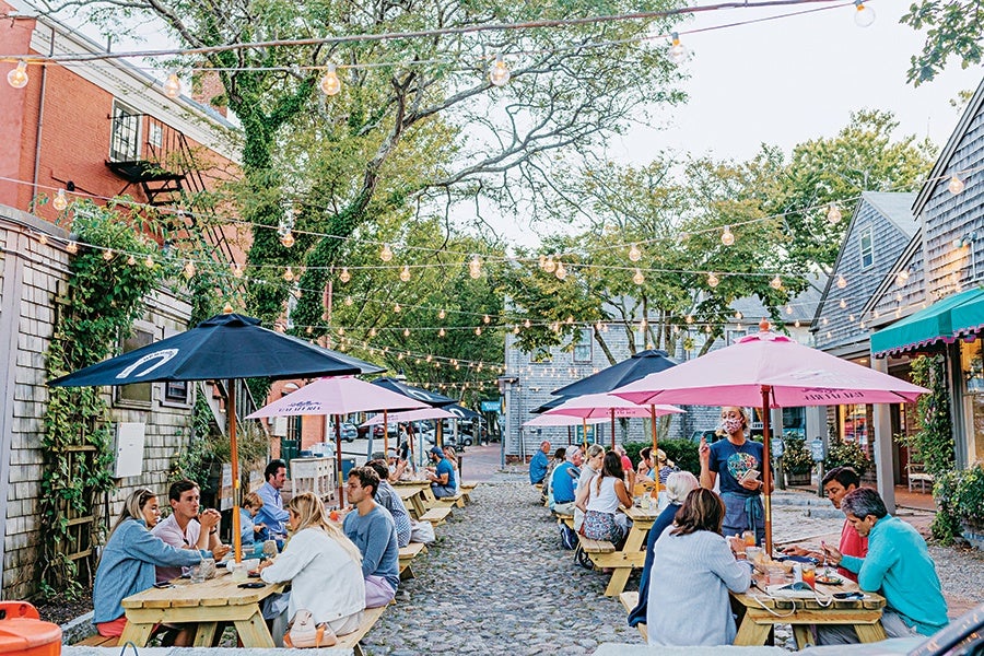Outdoor dining area on a cobblestone street with wooden picnic tables and umbrellas. People are seated at the tables enjoying food and drinks. String lights are hung overhead, creating a cozy atmosphere. Surrounding buildings have a mix of brick and wooden siding with greenery climbing the walls.