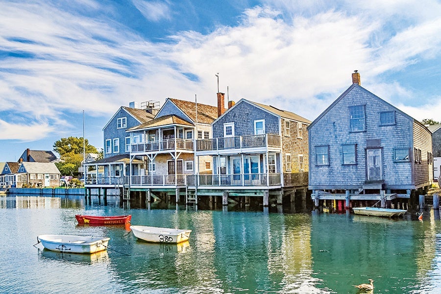Waterfront houses with wooden siding and porches on stilts above calm water, accompanied by three small boats floating nearby under a partly cloudy blue sky.