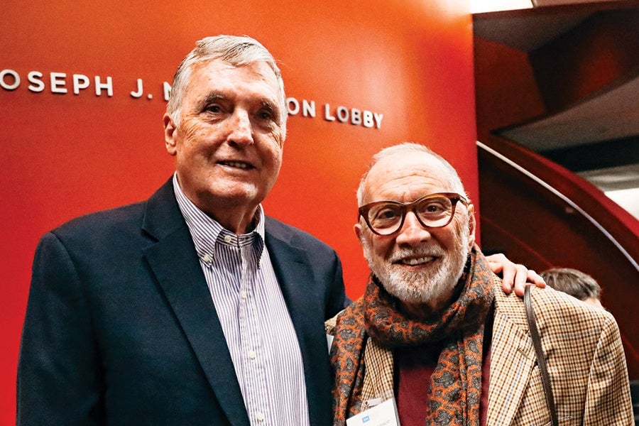 Two older men are standing closely together in front of an orange wall with partial text reading "OSEPH J. ... ON LOBBY." The man on the left has short gray hair and is wearing a dark blazer over a striped shirt. The man on the right has a white beard, glasses, and is dressed in a checkered jacket with a patterned scarf. Both men are smiling, and the man on the left has his hand on the other man's shoulder.