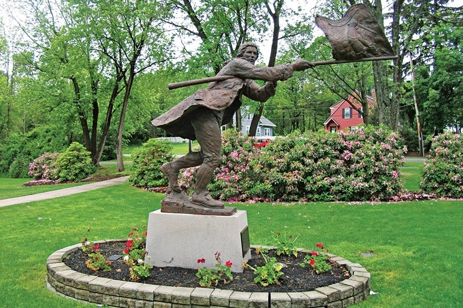 Bronze statue of a man in historical clothing holding a flag, positioned as if running or lunging forward, mounted on a stone pedestal surrounded by a circular flower bed with red flowers, set in a green park with trees and houses in the background.