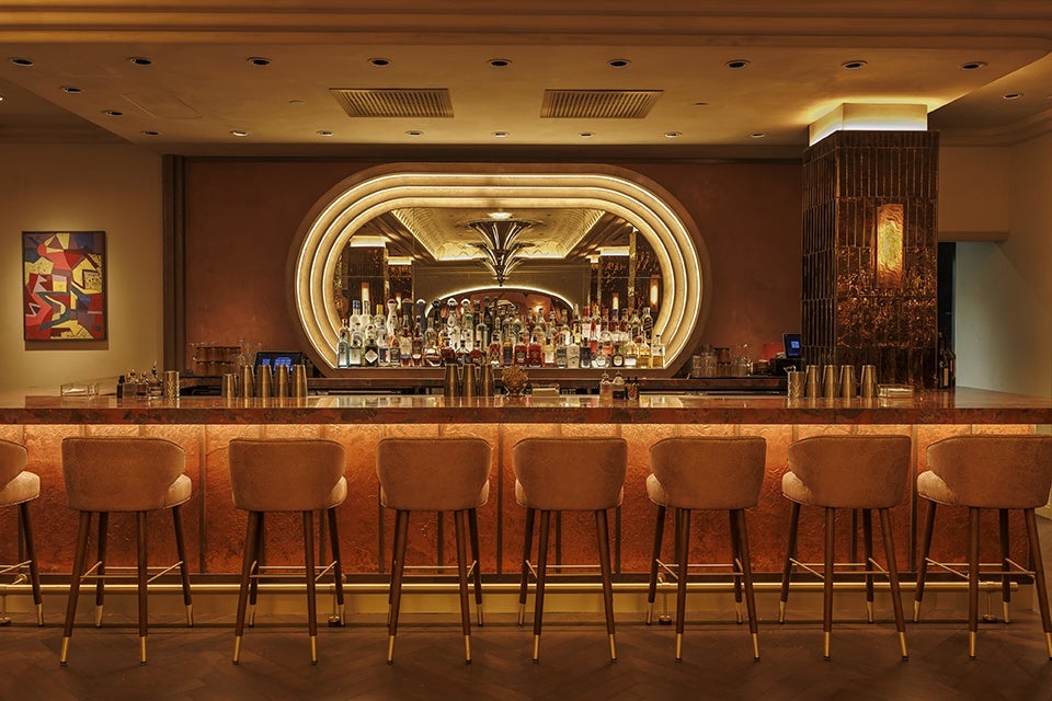 A stylish bar with a long counter illuminated by warm, amber lighting. Eight cushioned bar stools with wooden legs and gold accents are neatly lined up in front of the counter. Behind the bar, a large, oval-shaped mirror with layered lighting frames a wide selection of liquor bottles. The ceiling features recessed lighting, and a colorful abstract painting hangs on the left wall. The overall ambiance is cozy and elegant.