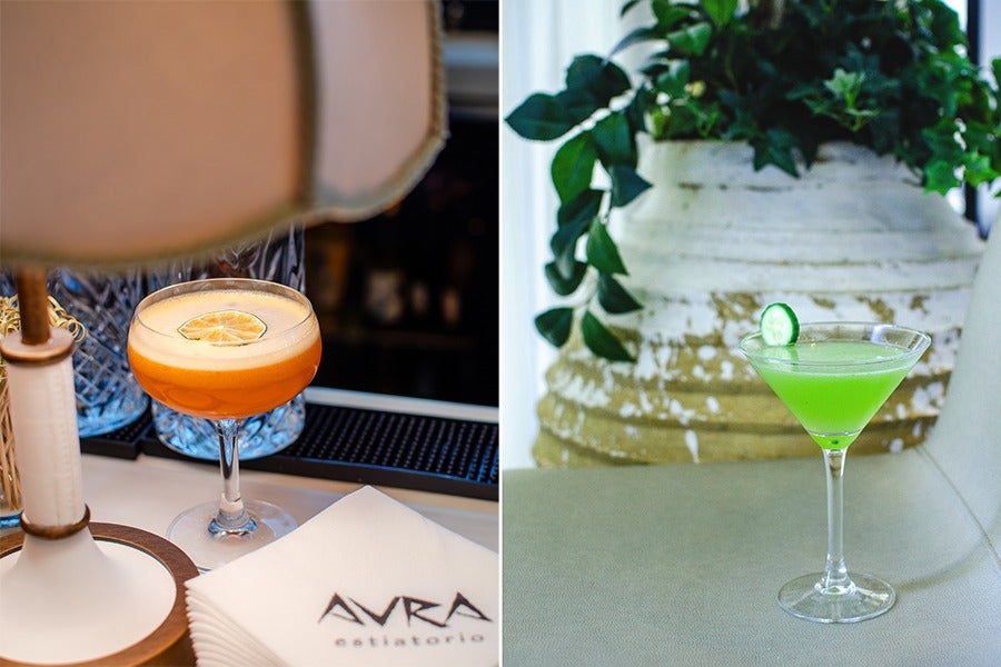 Two cocktails are shown side by side. The left cocktail is an orange drink in a coupe glass, garnished with a dried lemon slice, placed on a bar counter next to a lamp and napkins with "AVRA estiatario" printed on them. The right cocktail is a bright green drink in a martini glass, garnished with a cucumber slice, set on a light-colored surface with a large rustic planter and green foliage in the background.