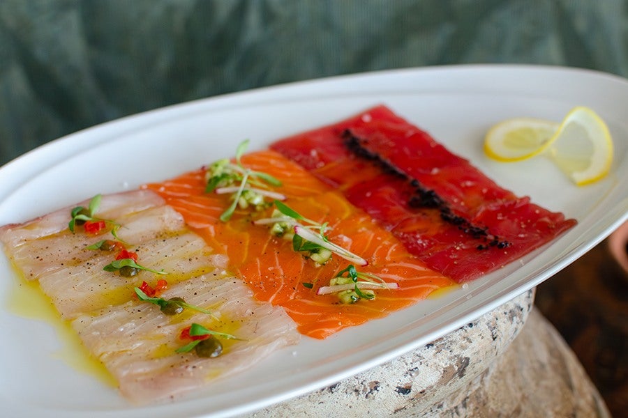 A white oval plate with three rows of thinly sliced fish carpaccio in different colors: white, orange, and red. Each row is garnished with small greens and seasonings, with a drizzle of olive oil on the white fish. A lemon twist is placed on the right side of the plate. The plate is set on a rustic stone surface.