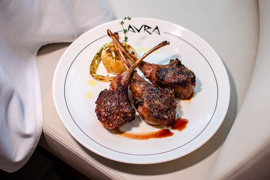Three grilled lamb chops seasoned with herbs are served on a white plate with a roasted garlic bulb and a drizzle of sauce, garnished with a sprig of thyme. The plate has the word "AVRA" printed on the rim.