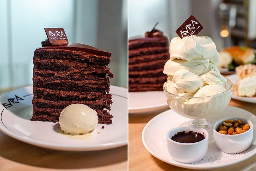 A tall slice of layered chocolate cake with a glossy chocolate topping and a small chocolate square labeled "AVRA estiatório," served on a white plate with a scoop of vanilla ice cream. Next to it, a glass bowl filled with a large swirl of vanilla soft serve topped with the same chocolate square, placed on a white plate alongside two small white bowls—one with chocolate sauce and the other with mixed nuts.