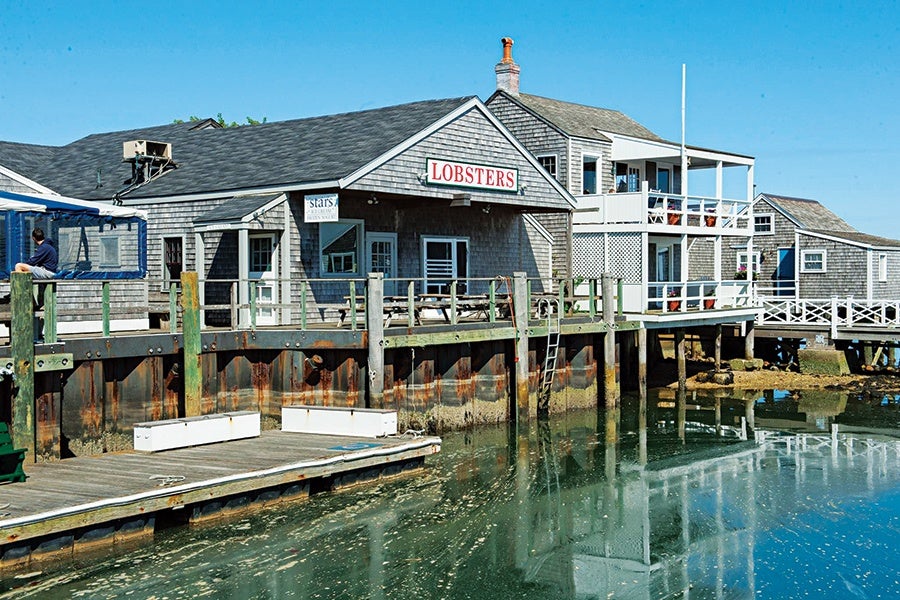 A wooden waterfront building with a sign reading "LOBSTERS" on the front. The building is situated on a dock above calm water, with weathered wooden pilings and a small ladder leading into the water. The structure has gray shingled siding and a pitched roof, with an adjacent building featuring balconies and white railings. A person is sitting on the dock to the left side of the image. The sky is clear and blue.