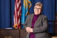 A woman with short gray hair and glasses is smiling, wearing a gray blazer over a purple turtleneck. She is standing with her hands clasped in front of her. Behind her are two flags, one of which is the American flag, and a nameplate on a wooden desk that reads "LIZ BREADON.