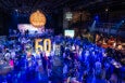 A large indoor event with a crowd of people dressed in semi-formal attire, illuminated by blue lighting. In the center, there is a prominent yellow "50" display on a white platform. Behind it, a stage features a car and a large circular sign that reads "50th Anniversary Best of Boston Soirée." Various booths and tables are set up around the room, with people mingling and engaging. The venue has a high ceiling with visible lighting rigs and a balcony area on the right side.