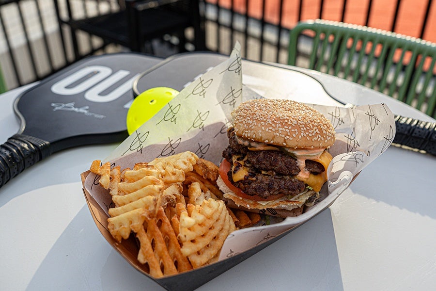 A sesame seed bun cheeseburger with two beef patties, melted cheddar cheese, lettuce, tomato, and sauce, served with a side of waffle fries in a paper-lined tray. A pickleball paddle and ball are visible in the background on a white table.