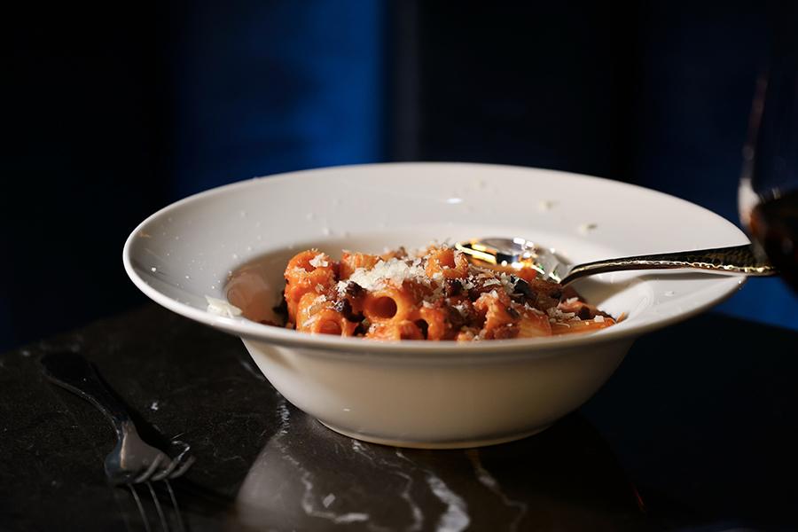 A white bowl filled with rigatoni pasta in a red tomato sauce, topped with grated cheese and small pieces of black olives. A spoon rests inside the bowl, and a fork is placed on the dark marble table beside it. The background is dark with a hint of blue.