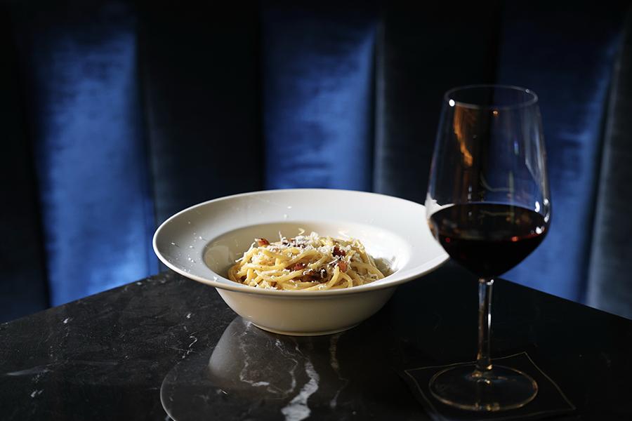A white bowl of spaghetti carbonara topped with grated cheese and bits of bacon, placed on a dark marble table. Next to the bowl is a glass of red wine. The background features a blue velvet upholstered surface.