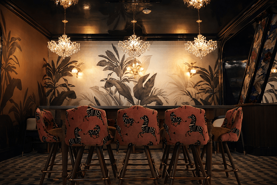A dining area with a long wooden table surrounded by pink upholstered chairs featuring black zebra patterns. The walls have large botanical murals in muted tones, and three ornate crystal chandeliers hang from the dark ceiling. The floor has a geometric patterned tile, and there are two small wall sconces with warm lighting on the mural wall. A large mirror with floral details is visible on the right side.