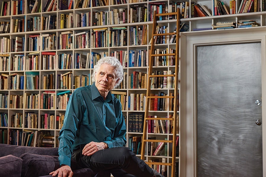 An elderly man with white hair wearing a teal button-up shirt and black pants is sitting on a dark purple couch. Behind him is a large bookshelf filled with numerous books, and a tall wooden ladder leans against the shelves. To the right, there is a metallic door with two round knobs.
