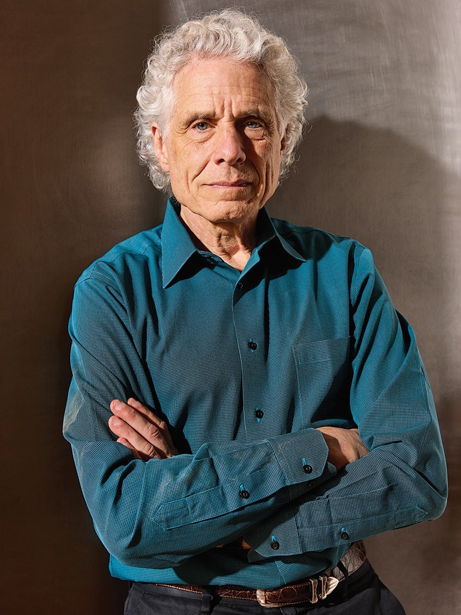 Older man with curly white hair wearing a teal button-up shirt and a brown belt, standing with arms crossed against a plain brown background.