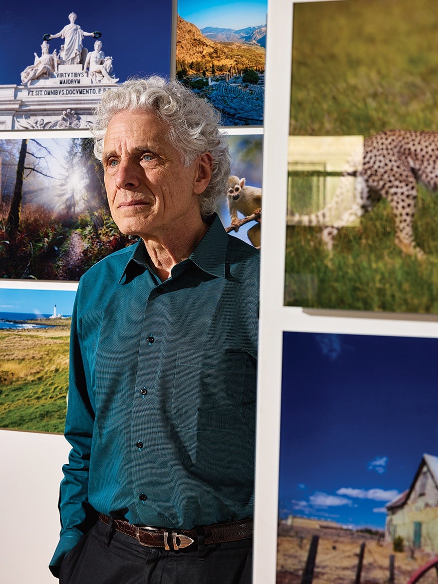 A man with curly gray hair wearing a teal button-up shirt and black pants stands with his hands in his pockets. He is positioned next to a wall displaying several colorful photographs, including images of a statue, a mountainous landscape, a forest path, a small animal on a branch, a cheetah in grass, a lighthouse by the sea, and a rural house under a blue sky. The man looks thoughtfully into the distance.