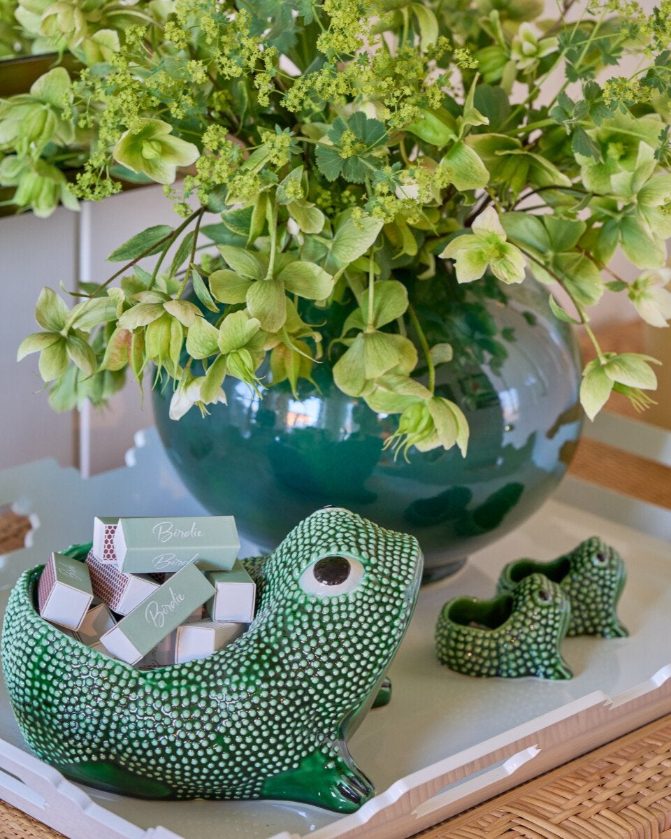 A green ceramic frog-shaped container filled with small rectangular boxes labeled "Birdie" sits on a white tray. Next to it are two smaller matching frog-shaped ceramic pieces. Behind the tray is a large, round, dark green vase filled with green flowers and foliage. The setting appears to be on a wicker surface.