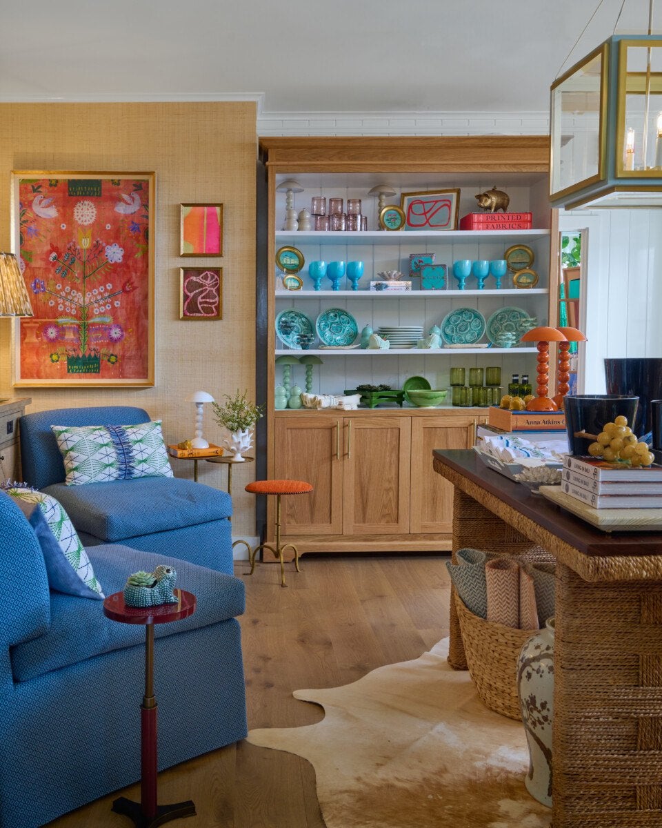 Cozy living room featuring blue upholstered seating with patterned pillows, a wooden cabinet with open shelves displaying turquoise and green dishware, glassware, and decorative items. The walls have a textured beige finish with colorful framed artwork. A woven table with books, black planters, and a basket with rolled textiles sits on a light cowhide rug. Warm wood flooring and a large geometric pendant light complete the space.