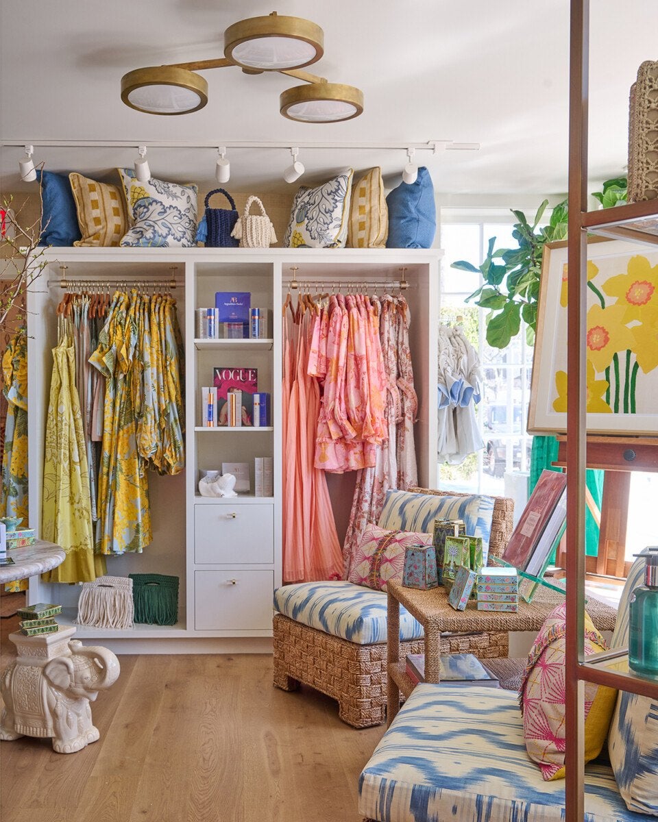 A bright, stylish boutique interior featuring a white shelving unit with hanging colorful dresses in yellow, pink, and floral patterns. The top of the shelving unit is decorated with patterned pillows and small woven handbags. In front of the shelves, there are woven rattan chairs with blue and white patterned cushions and colorful throw pillows. A small woven table between the chairs displays various boxed products. The space has a wooden floor, a modern gold ceiling light fixture, and a large window letting in natural light. A framed floral artwork and green plants add to the cheerful, inviting atmosphere.