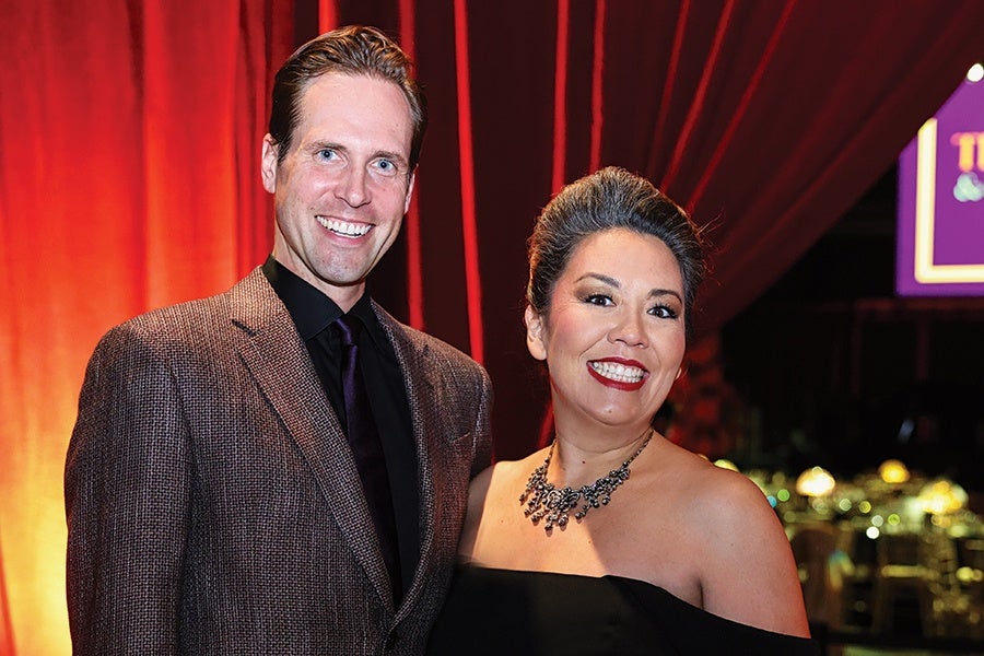 A man and a woman smiling and posing together in front of red curtains. The man is wearing a brown textured suit jacket with a black shirt and dark tie. The woman is wearing an off-the-shoulder black dress and a statement necklace with dark stones. The background includes blurred lights and a purple sign.