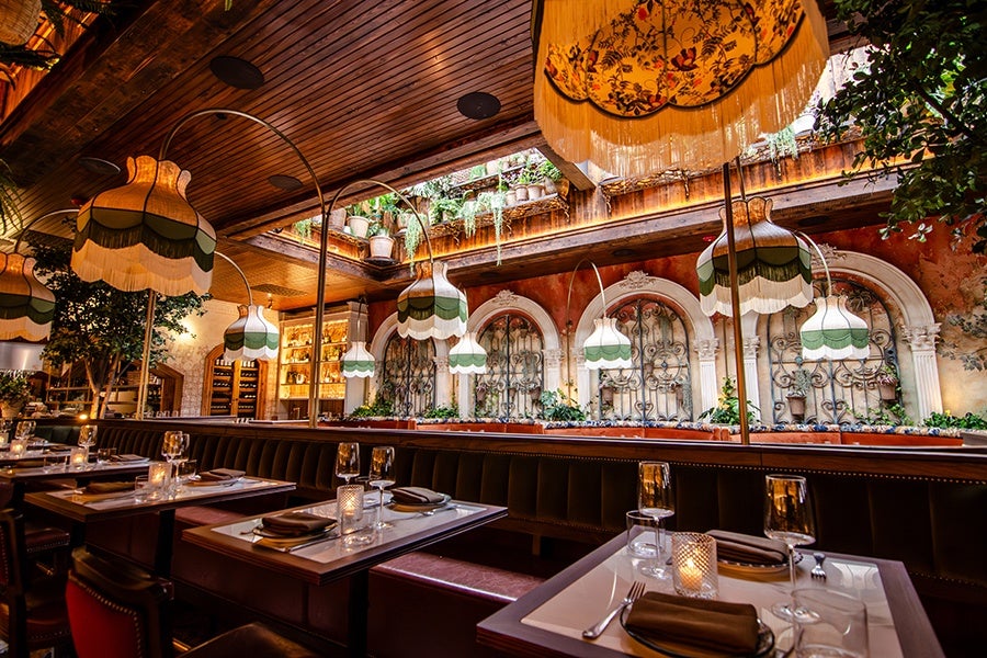 Cozy restaurant interior featuring dark leather banquette seating and wooden tables set with glassware, plates, and napkins. The ceiling is wooden with large, vintage-style hanging lampshades in cream, green, and orange tones. The walls have ornate arched windows with decorative wrought iron and potted plants, creating a warm and inviting atmosphere.