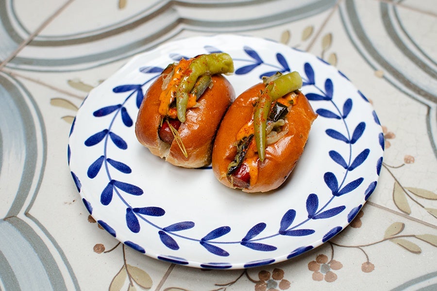 Two small hot dogs with grilled green chili peppers and a drizzle of orange sauce on top, served on a white plate with a blue leaf pattern. The plate is placed on a tiled surface with a floral and circular design.