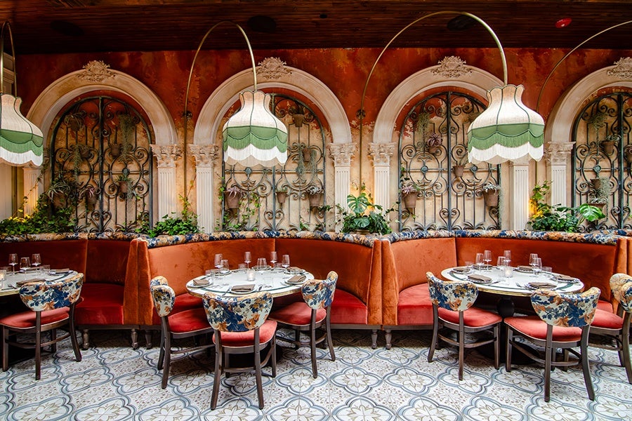 A cozy restaurant seating area features curved orange velvet banquettes paired with floral-patterned chairs with red cushions. Round tables are set with glassware, plates, and napkins. The background showcases ornate arched wall niches with decorative wrought iron grilles and climbing plants. Large, vintage-style floor lamps with scalloped green and white shades arch over the tables. The floor is tiled with a patterned design in soft colors.