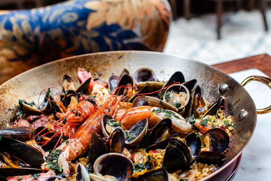 A large pan of seafood paella featuring shrimp, mussels, clams, and rice garnished with herbs. The pan has a brass handle and is set on a table with a patterned chair in the background.
