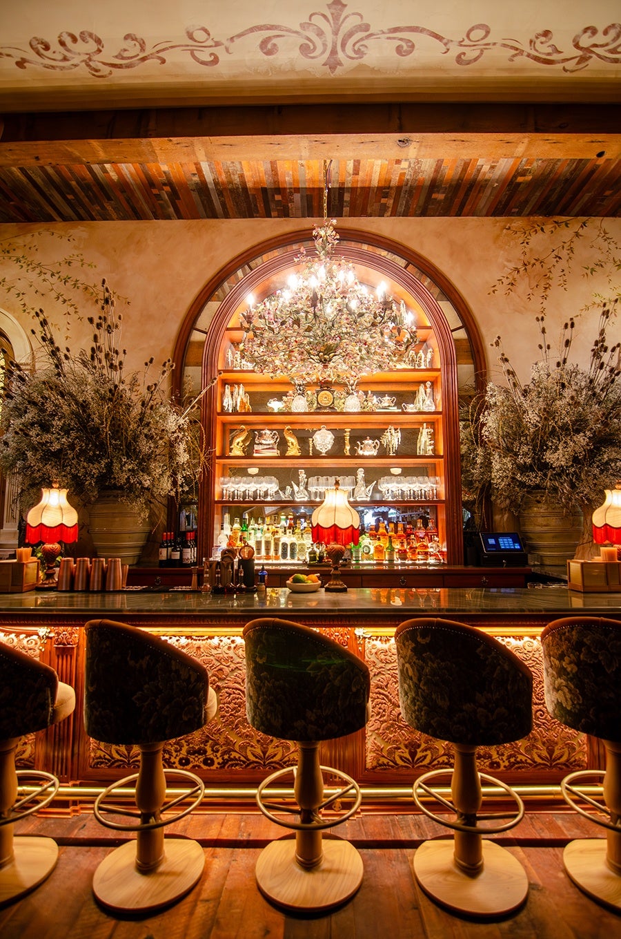 A luxurious bar interior featuring a polished wooden counter with ornate carved panels illuminated by warm lighting underneath. Five upholstered bar stools with wooden bases are lined up in front. Behind the bar, a large arched mirror and wooden shelves display an array of liquor bottles, glassware, and decorative items. A grand, ornate chandelier with floral details hangs above, flanked by large vases filled with dried floral arrangements. Two vintage-style lamps with red and cream shades add to the warm, elegant ambiance. The ceiling has wooden beams and decorative painted trim.