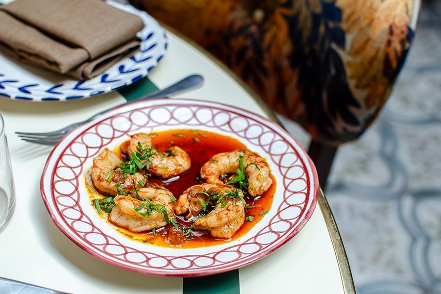 A plate with five cooked shrimp in a reddish sauce, garnished with chopped green herbs, on a white table. A fork and a glass are nearby, and a plate with a folded brown napkin is partially visible in the background.