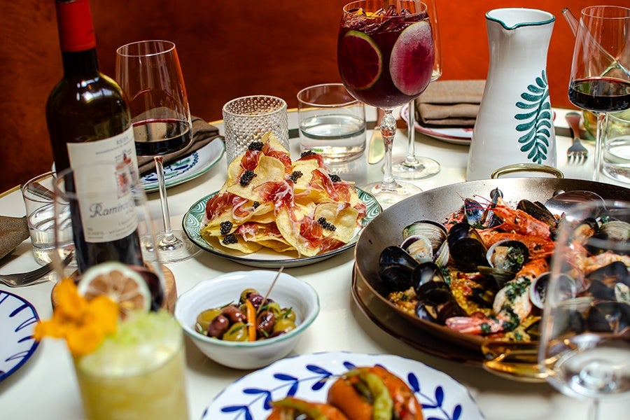 A table set with a variety of dishes and drinks, including a large pan of seafood paella with shrimp, mussels, and clams, a plate of nachos topped with cured meat and black caviar, a bowl of mixed olives, a bottle of red wine, glasses of red wine, a glass of sangria with fruit slices, and a ceramic pitcher with a green leaf design. The tableware features white plates with blue patterns.