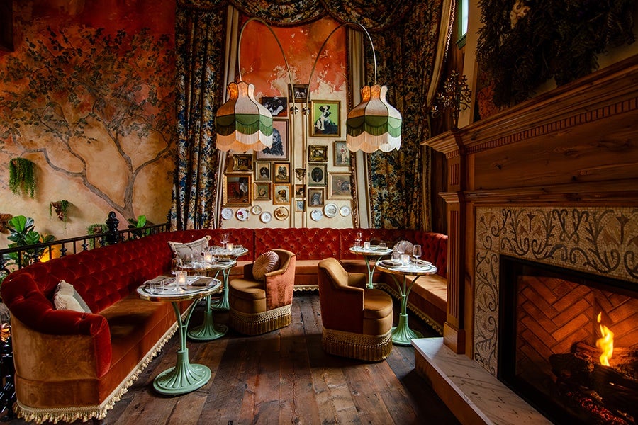 Cozy vintage-style seating area with a red velvet tufted sectional sofa and two matching armchairs with fringe trim, arranged around small round tables with glass tops. The space features a wooden floor, a lit fireplace with ornate tile surround, and walls adorned with framed pictures and decorative plates. Two large, scalloped lampshades hang from a curved floor lamp above the seating. The background includes floral-patterned curtains and a mural of a tree on a warm-toned wall.
