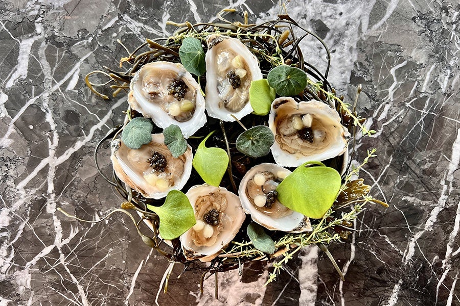Six oysters served on a bed of seaweed and garnished with small green leaves, presented on a dark marble surface. Each oyster contains small dollops of a creamy topping and a black garnish, possibly caviar.