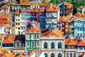 A dense cluster of colorful buildings with orange-tiled roofs. The buildings vary in height and style, featuring multiple windows, balconies, and some with solar panels on the roofs. The facades are painted in vibrant colors including green, blue, yellow, red, and white. The architecture has a European feel with ornate window frames and decorative elements.