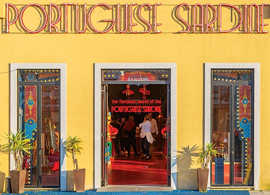 A bright yellow building facade with three doorways, the central one open showing people inside. Above the entrance, a large neon sign reads "PORTUGUESE SARDINE" in red letters. The two side doors have colorful decorative panels with blue, red, and gold designs. Two potted plants flank each side of the entrance.