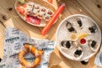 A wooden table displays a plate of sushi rolls with pickled ginger, wasabi, and soy sauce packets, accompanied by red chopsticks. Next to it is a round plate of oysters on ice, each topped with black caviar, garnished with a lemon wedge and small containers of sauce. A soft pretzel sprinkled with coarse salt rests on branded paper nearby.