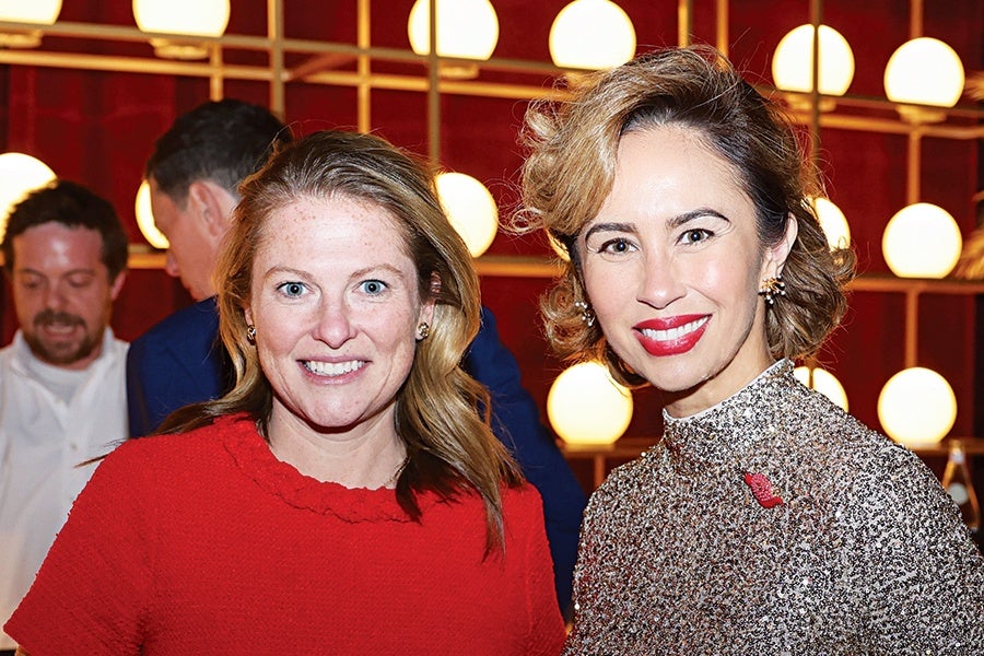 Two women smiling at the camera in a warmly lit indoor setting. The woman on the left has light skin, long blonde hair, and is wearing a red textured top. The woman on the right has light skin, short wavy light brown hair, red lipstick, and is wearing a sparkly silver top with a small red pin. The background features a red wall with a grid of round white lights.