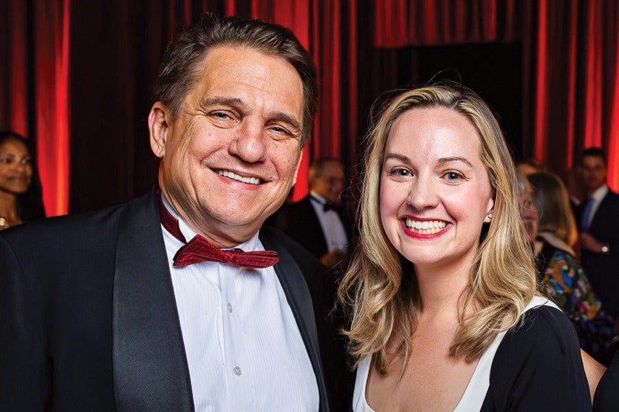 A man and a woman smiling at the camera in a formal setting. The man is wearing a black tuxedo with a white shirt and a red bow tie, while the woman has blonde hair and is wearing a black and white dress. The background features red curtains and other people dressed in formal attire.