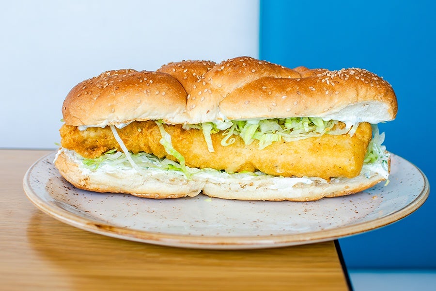 A sandwich with a sesame seed bun filled with a large piece of breaded fried fish, shredded lettuce, and a spread of mayonnaise or tartar sauce, served on a speckled ceramic plate.
